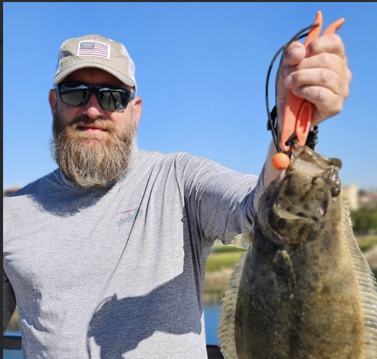 Man with a beard and sunglasses holding up a large flat fish with orange fish gripper, smiling outdoors.